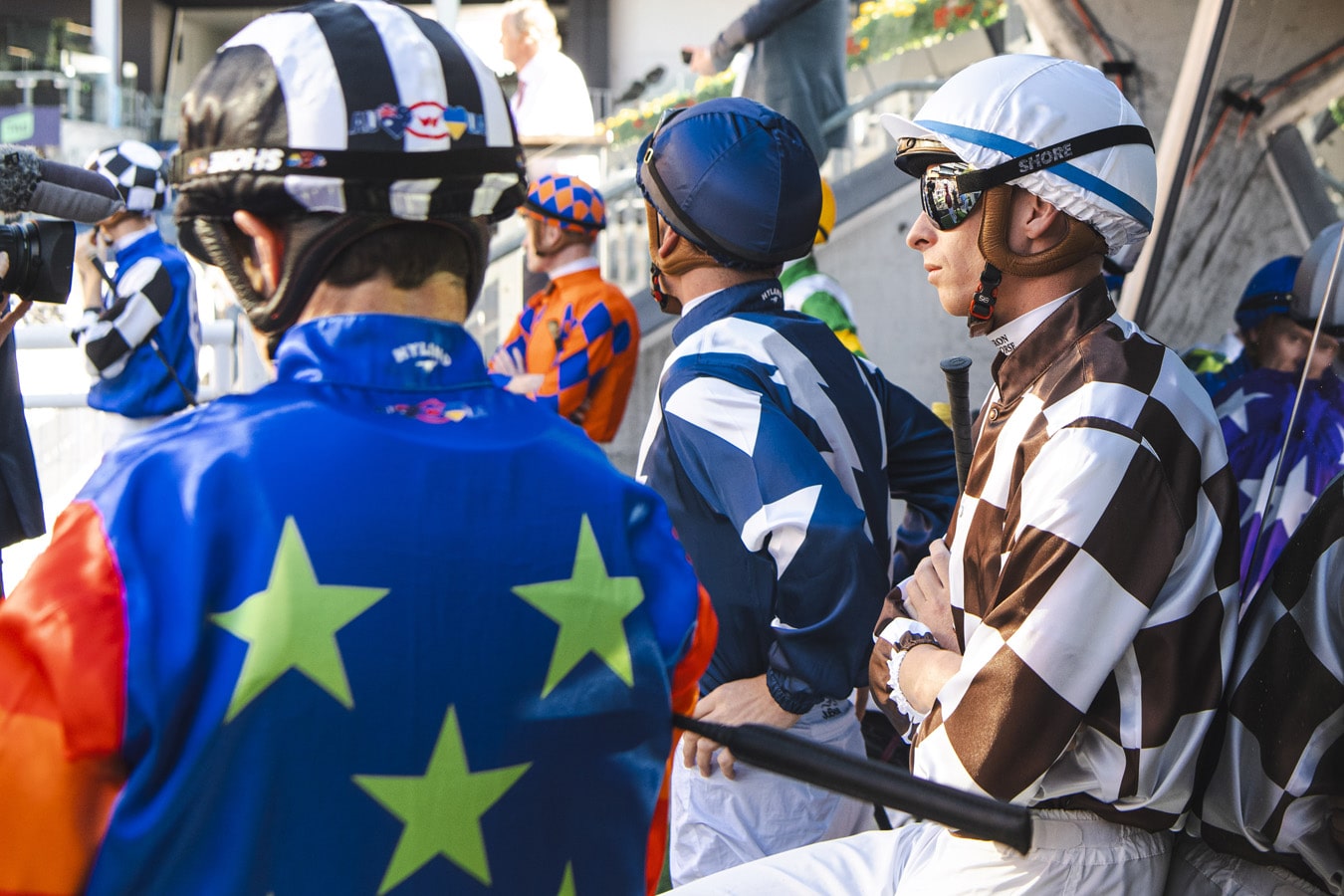 A group of jockeys wearing colours stand around waiting at a racecourse
