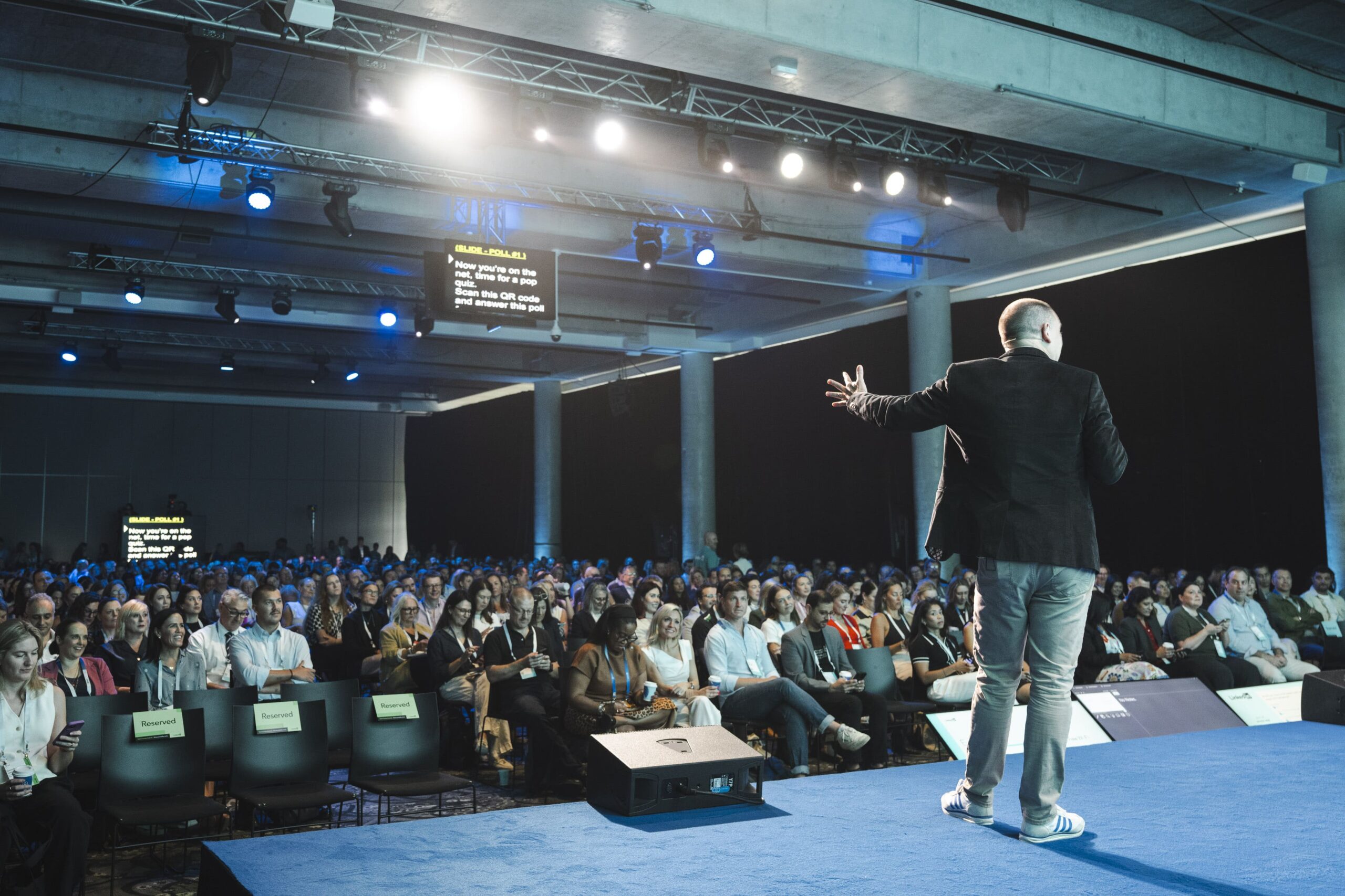A presenter is shown on stage in a crowded conference setting