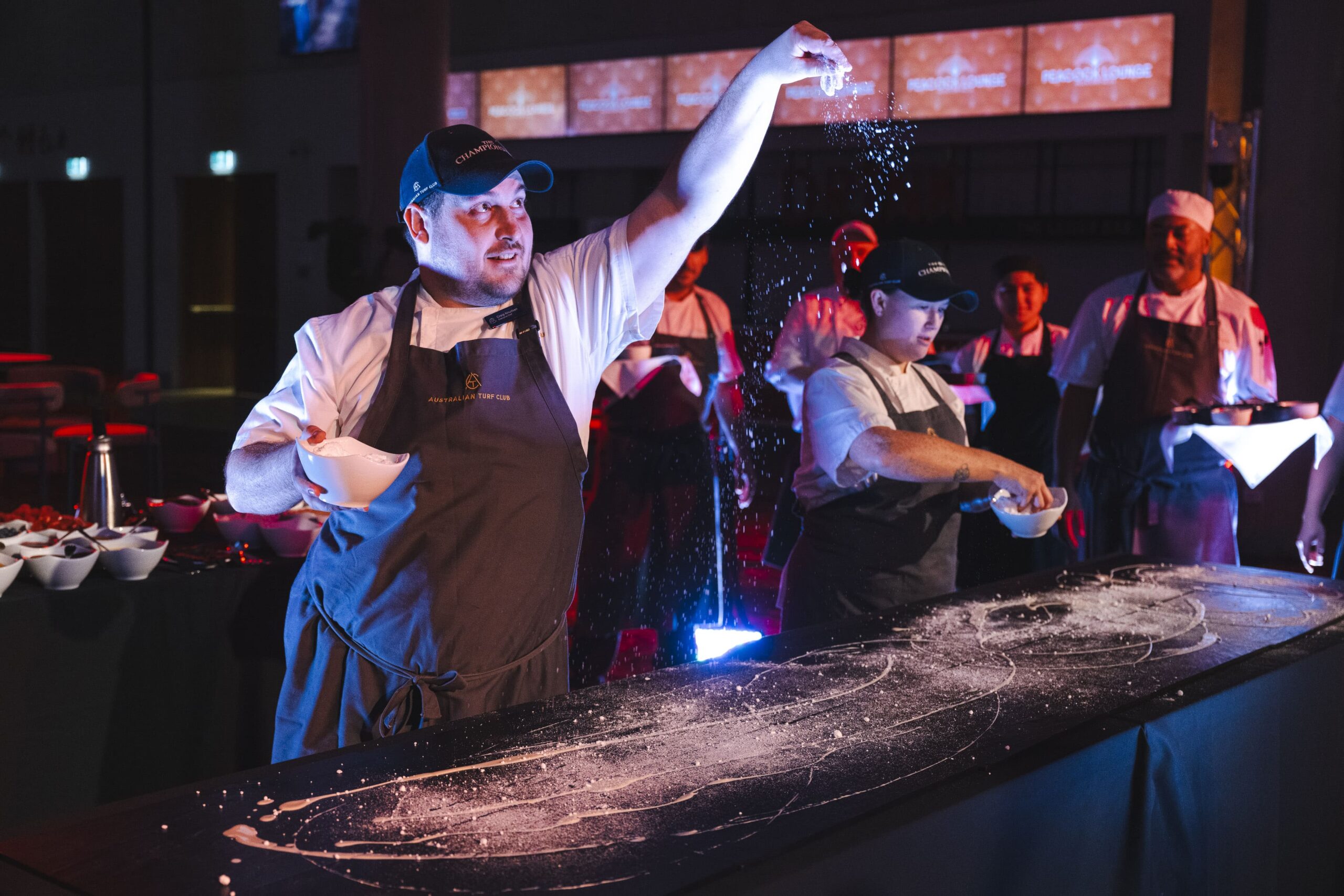 Chefs are shown sprinkling ingredients onto a bench in front of them in a culinary performance